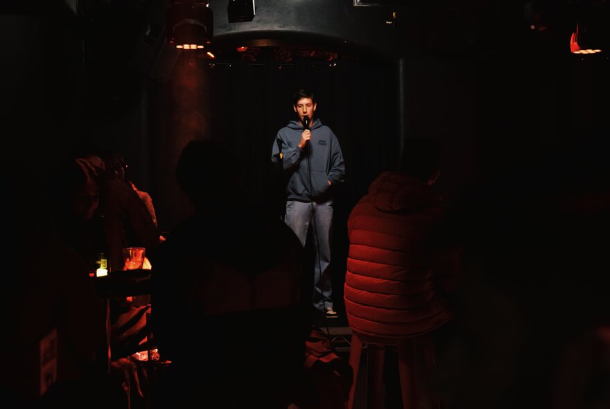 A comedian holding a microphone on stage in a darkened venue.