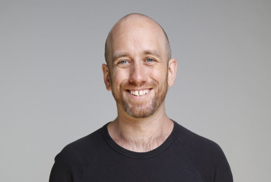 Comedian Danny McGinlay smiling in front of a white backdrop, wearing a black t-shirt.