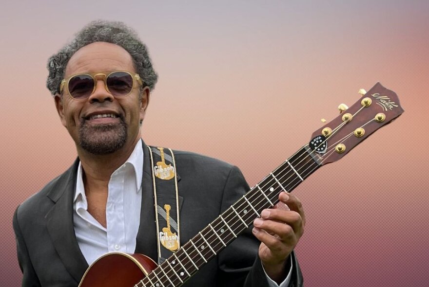Singer and guitarist Little Paul, with sunglasses on, guitar in hand, smiling.