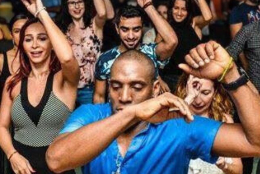 A crowd of people dancing to Cuban music, a man in a blue shirt leading from the front.