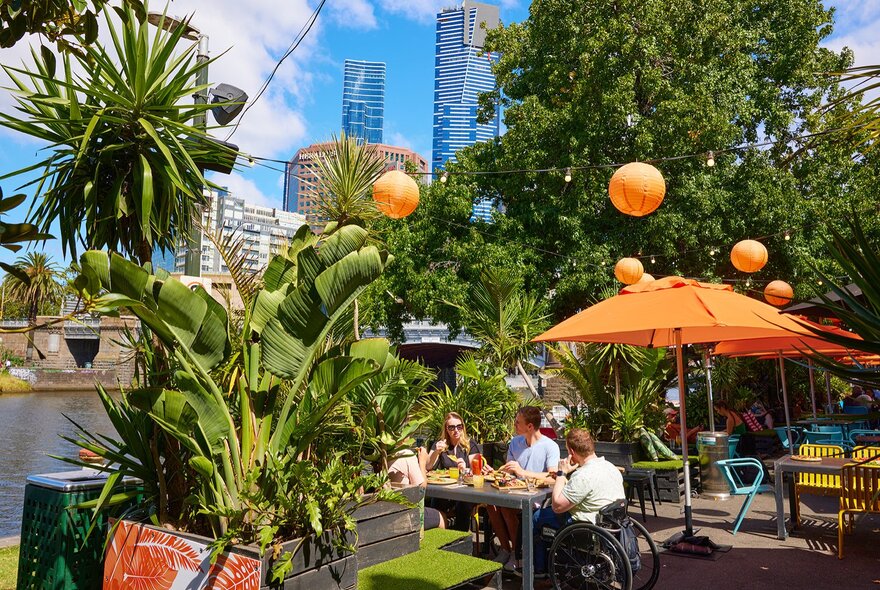 A group dining in a sunny outdoor bar by the river with orange lanterns and umbrellas.