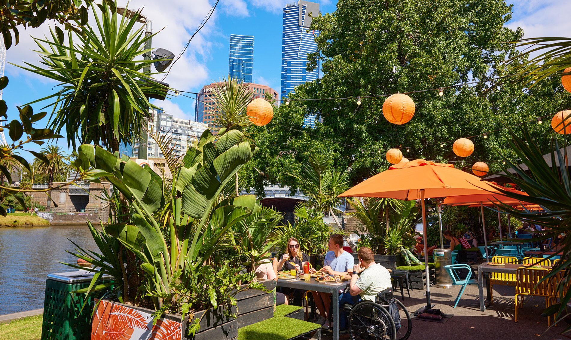 A group dining in a sunny outdoor bar by the river with orange lanterns and umbrellas.