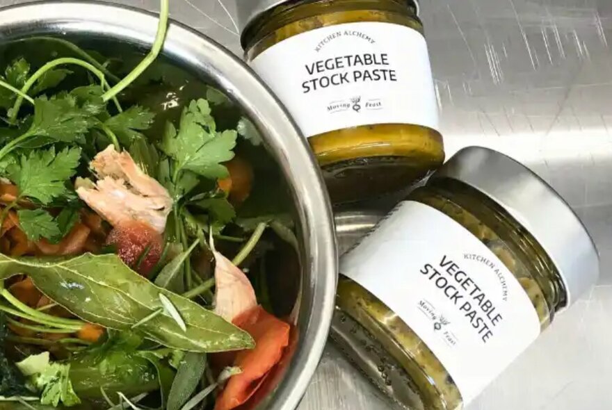 A bowl of raw vegetables and herbs, and two small jars of home-made vegetable stock paste next to it.