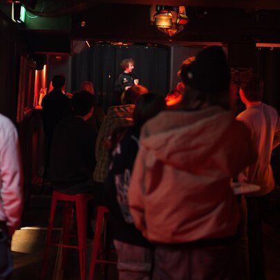 A dark image of a comedian on stage seen from behind a standing audience.