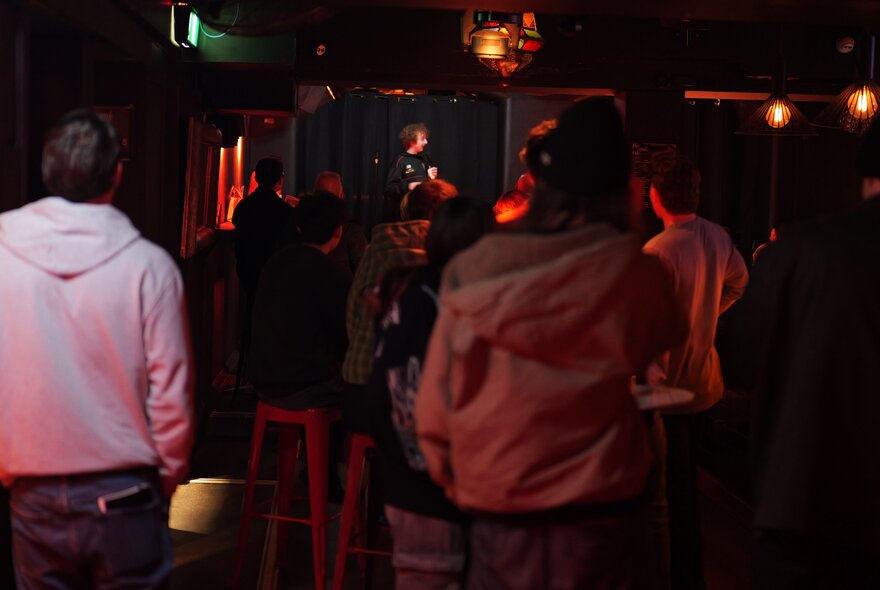 A dark image of a comedian on stage seen from behind a standing audience.