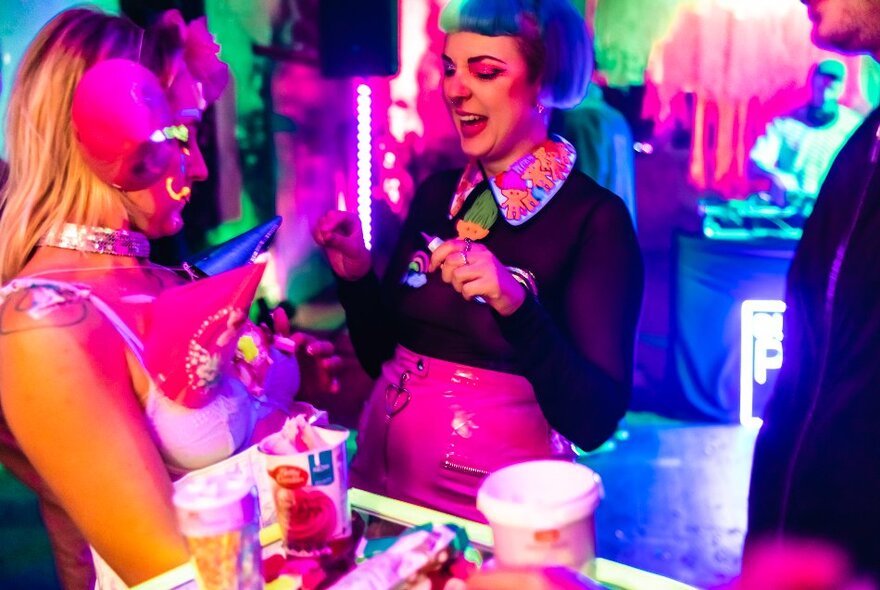 Two women wearing neon outfits in a bar setting with neon lights all around them, celebrating a birthday.