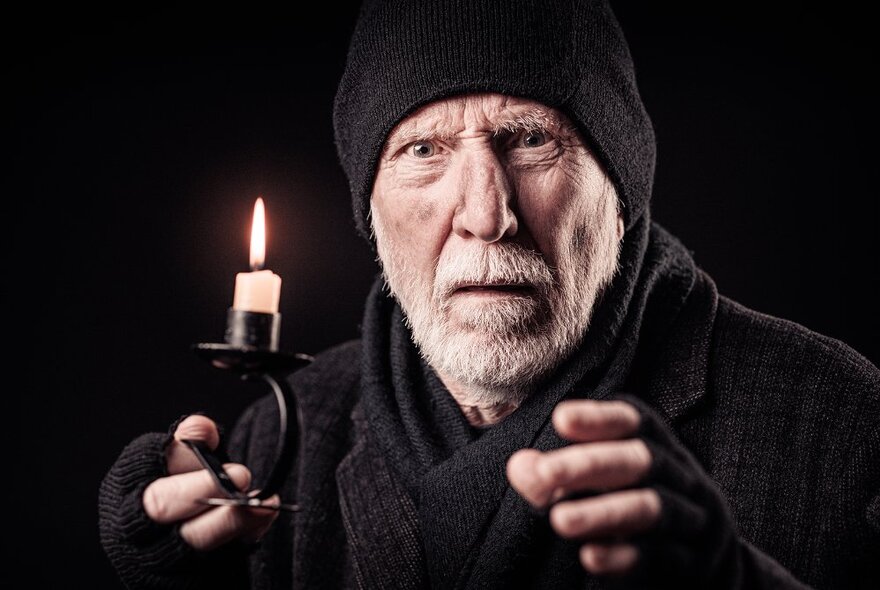 An older man wearing a black beanie, scarf, jacket and fingerless gloves, holding a short black candelabra with a stub of a lit candle.