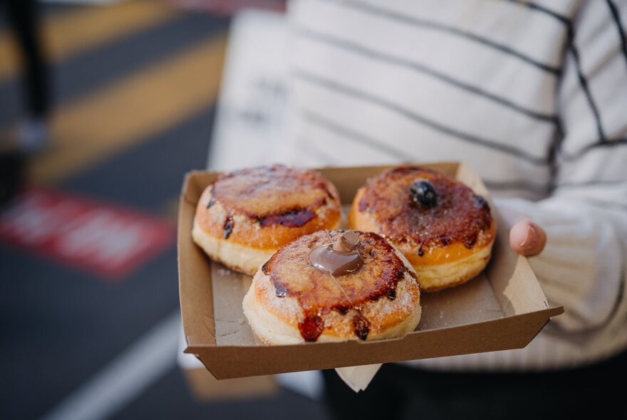 A hand holding a cardboard plate with three flavoured donuts.