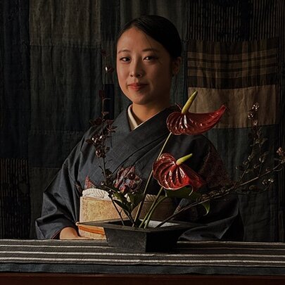Woman wearing a kimono, seated behind a striped table covering with stylised ikebana arrangement.