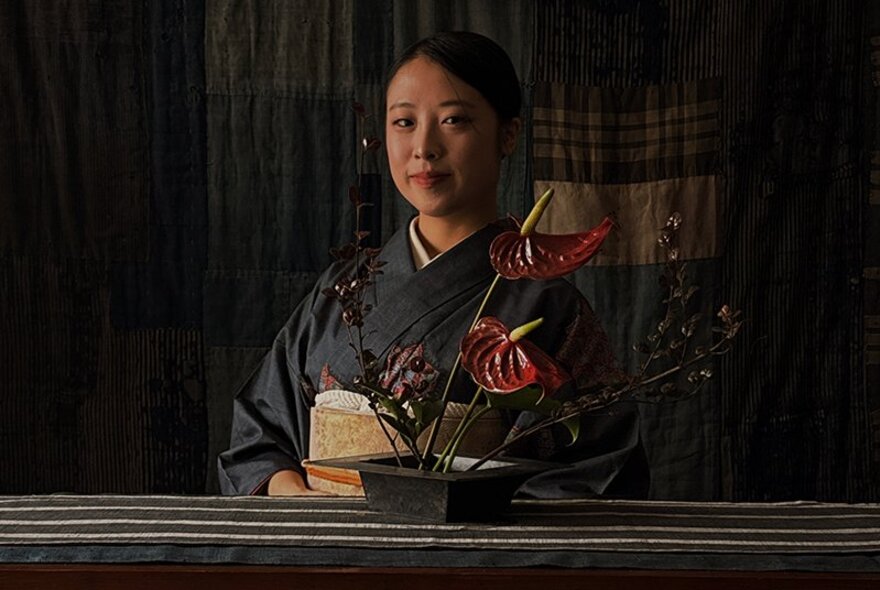 Woman wearing a kimono, seated behind a striped table covering with stylised ikebana arrangement.