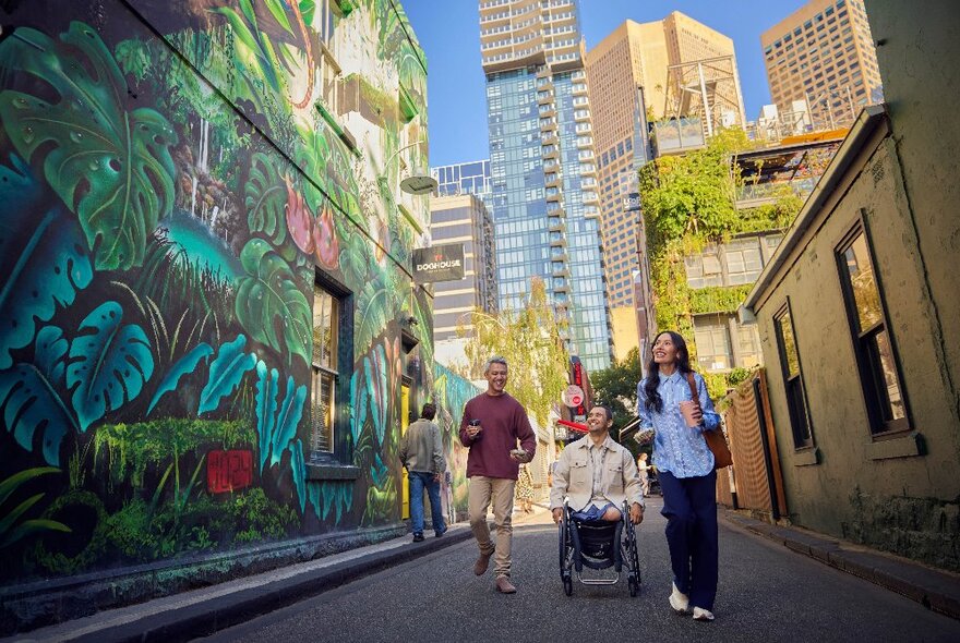 People including a man in a wheelchair heading down a laneway past a large tropical-themed wall mural with city buildings in the background.