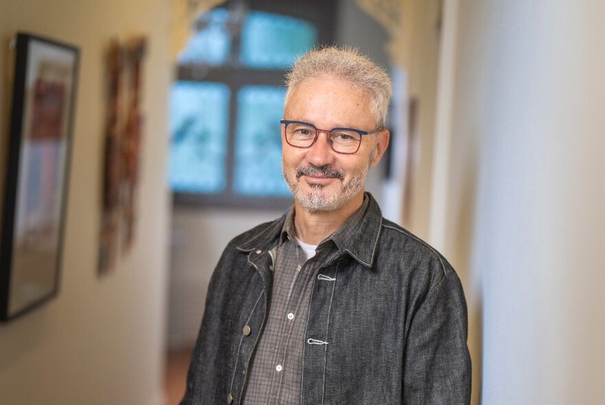 Author Josh Bornstein standing in a room with pictures on the wall, wearing a dark grey denim jacket, with short greying hair and a neat beard, wearing glasses and with a slight smile on his face.