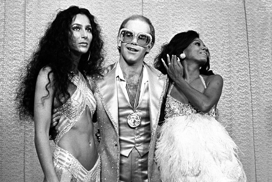 Elton John in the 1970s wearing large spectacles and satin suit with Cher wearing Bob Mackie midriff-revealing strappy gown and Diana Ross wearing feather and sequin gown.