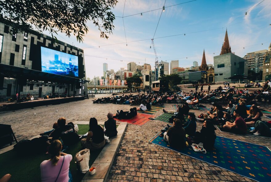 A crowd of people seated on picnic rugs and deck chairs outside at Fed Square, watching an open air film on a large screen.