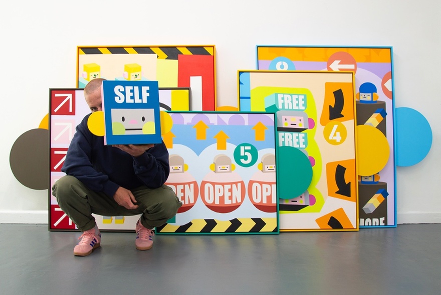 A collection of framed Conrad Square artworks leaning against a white wall, with the artist crouching in front of them holding a small artwork in front of his face.