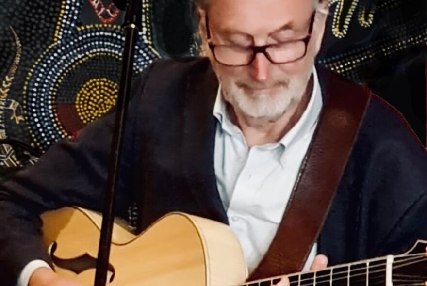 Man playing guitar at a microphone, with Indigenous pattern in the background.