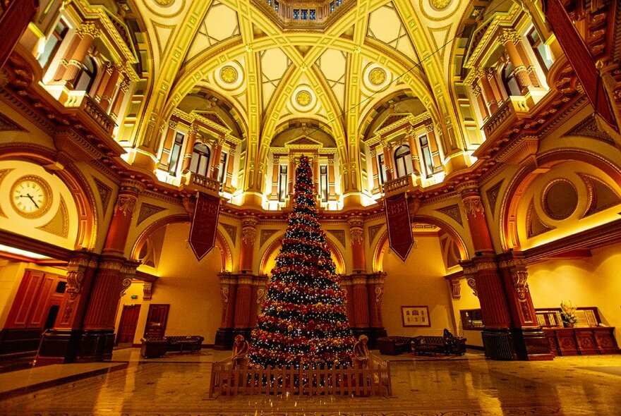 A towering Christmas tree with red decorations stands in the foyer of a baroque building foyer bathed in gold light.