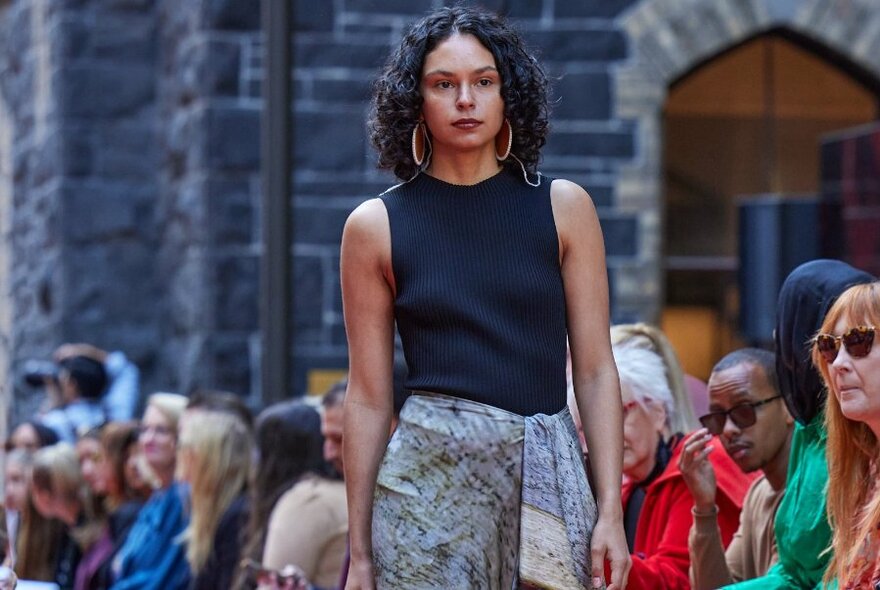 Model walking down an outdoor runway, with seated onlookers, wearing a black tank top and a light coloured wrap-around skirt.