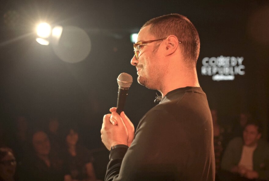 A comedian in profile on stage in front of an audience, holding a microphone close to his mouth.