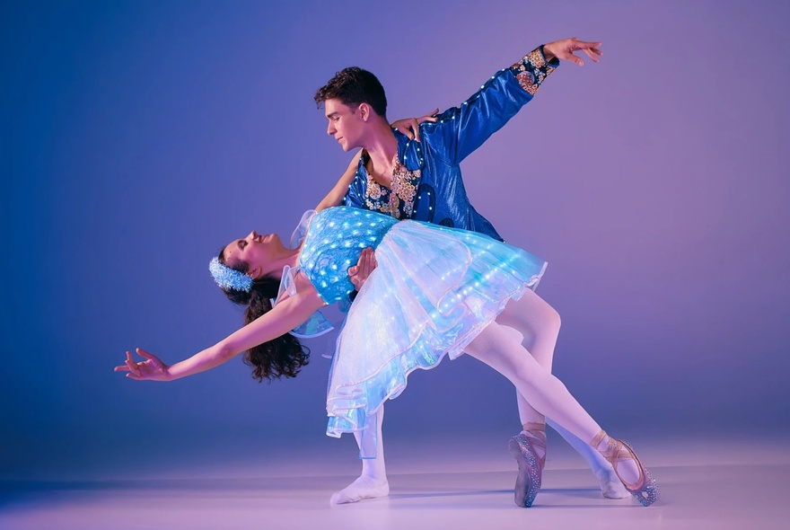 Two ballet dancers, one male and one female, engaged in a dance together, both dressed in blue-toned glow-in-the-dark costumes.