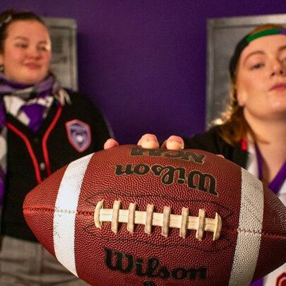 Two young women in school uniform in a purple room, one holding forward an American football. 