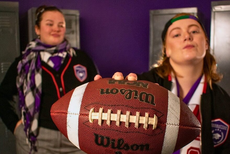 Two young women in school uniform in a purple room, one holding forward an American football. 