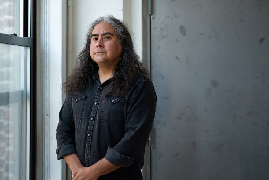 Composer and musician Raven Chacon, wearing a dark coloured shirt, leaning against a white wall near a window, his head turned and looking at the viewer.