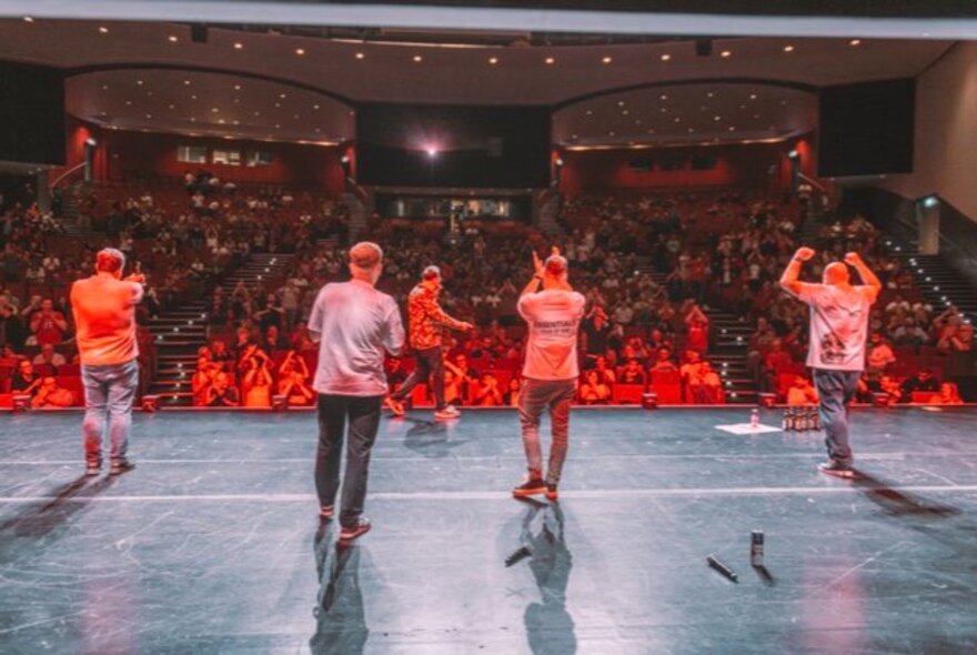 View of a seated audience in a large theatre from the back of a stage, with five men on stage seen from behind clapping and cheering.