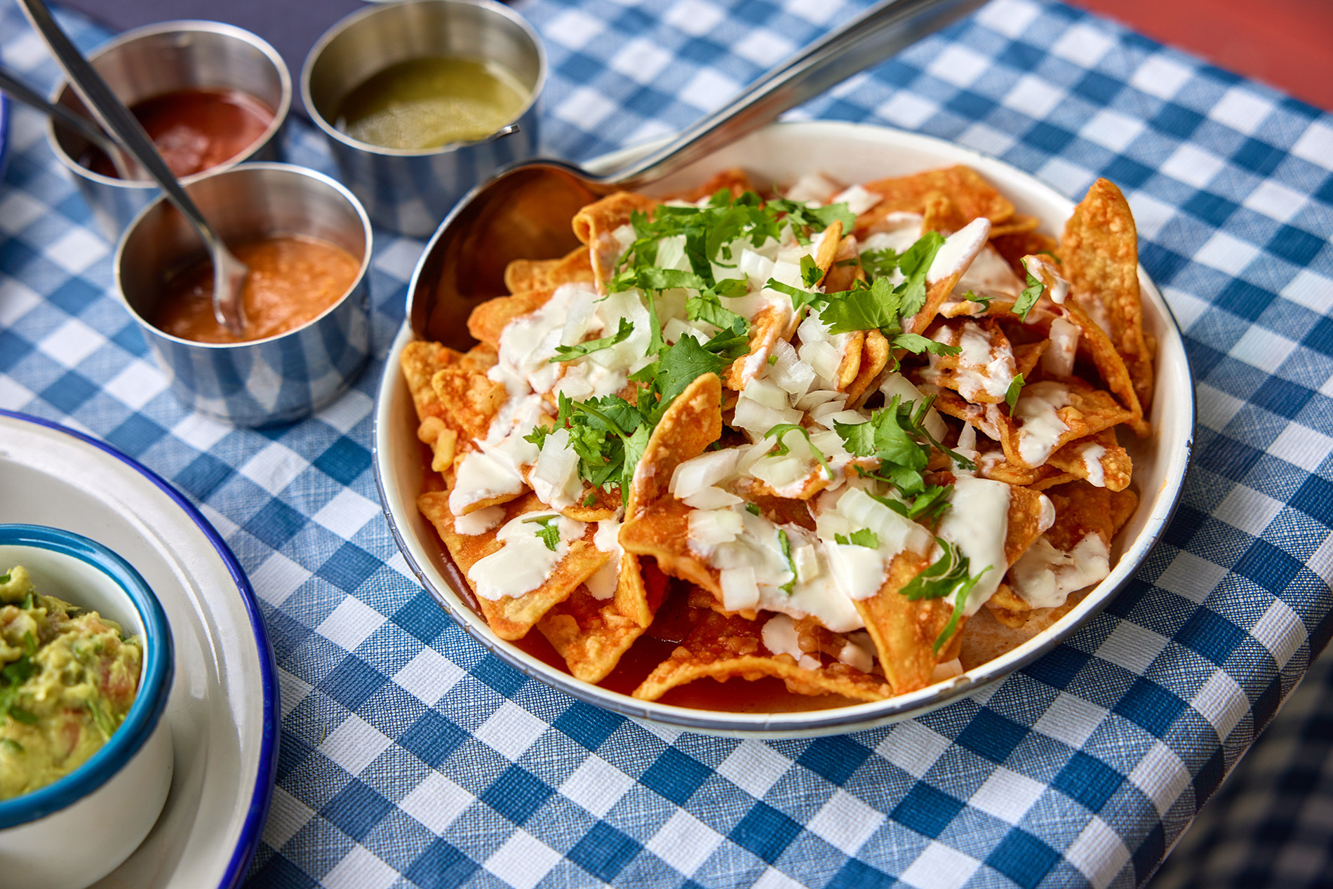 A bowl of nachos with sauces on the side on a blue checked tablecloth.