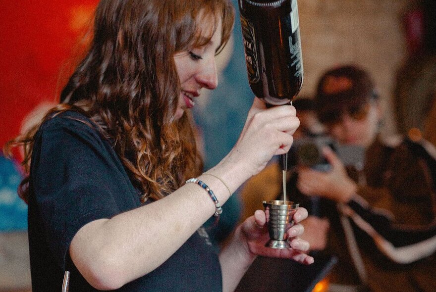 A woman with long curly hair pouring alcohol into a jigger.