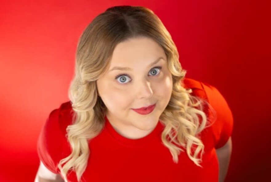 Stand-up comedian Mish Wittrup in a red dress with her face looking up, her curled blonde hair falling past her shoulders.