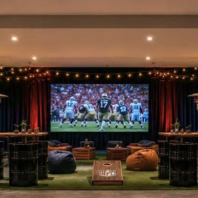 An underground carpark transformed into a tailgate party with a projector screen showing NFL, a green carpet with bean bags and bar tables with barrel seats.