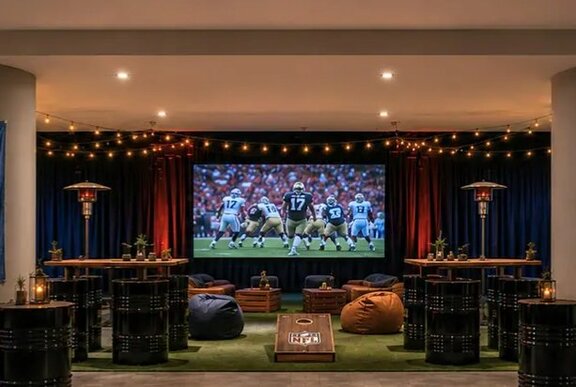 An underground carpark transformed into a tailgate party with a projector screen showing NFL, a green carpet with bean bags and bar tables with barrel seats.