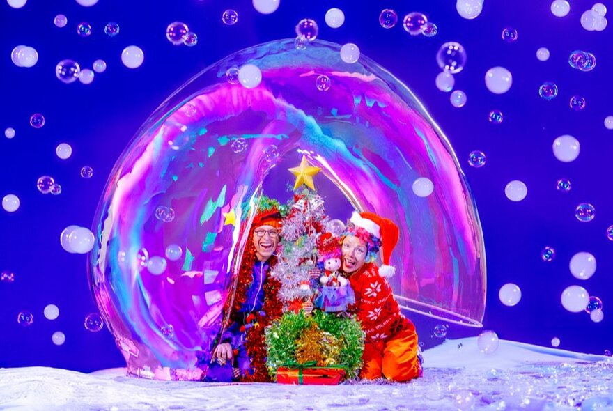 Two performers dressed in Christmas costumes, smiling next to a Christmas tree, encased in a giant bubble and surrounded by bubbles, against a blue background.