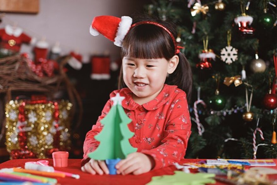 A little girl with a Santa hat headband holding a craft Christmas tree on a table in front of a real Christmas tree. 