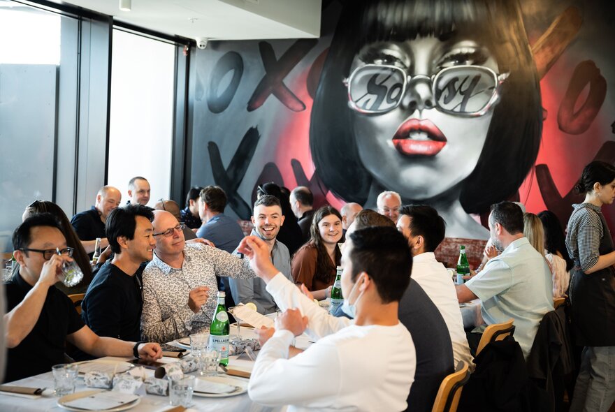 A packed dining room with large tables of families and friends enjoying a meal with a large mural of a woman's face looking down on them. 
