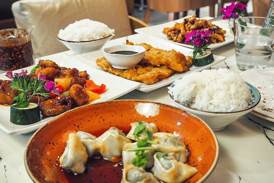 Array of dishes on a table including dumplings in orange dish, rice and stir-fries.