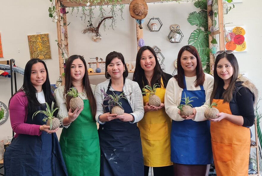 Six class participants in coloured aprons holding up kokedama balls. 