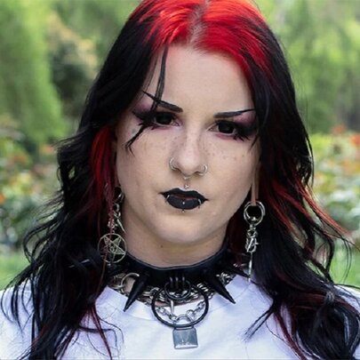 A person styled with alternative gothic fashion, featuring black hair with red highlights, dramatic dark makeup, and a spiked choker necklace.