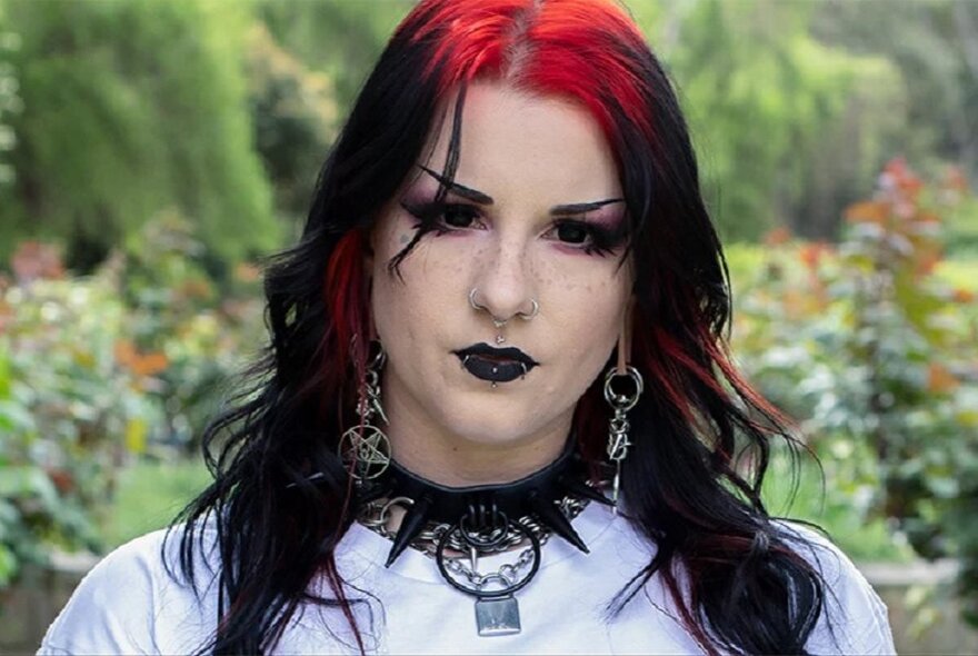 A person styled with alternative gothic fashion, featuring black hair with red highlights, dramatic dark makeup, and a spiked choker necklace.