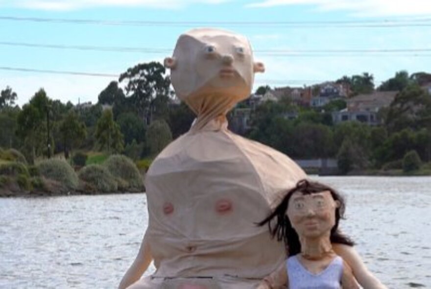 Woman wearing a mask standing with a large puppet sculpture of a naked baby on a waterway.