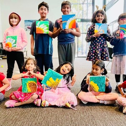 A standing and seated row of children in a large bright room, all holding up artworks they've created.