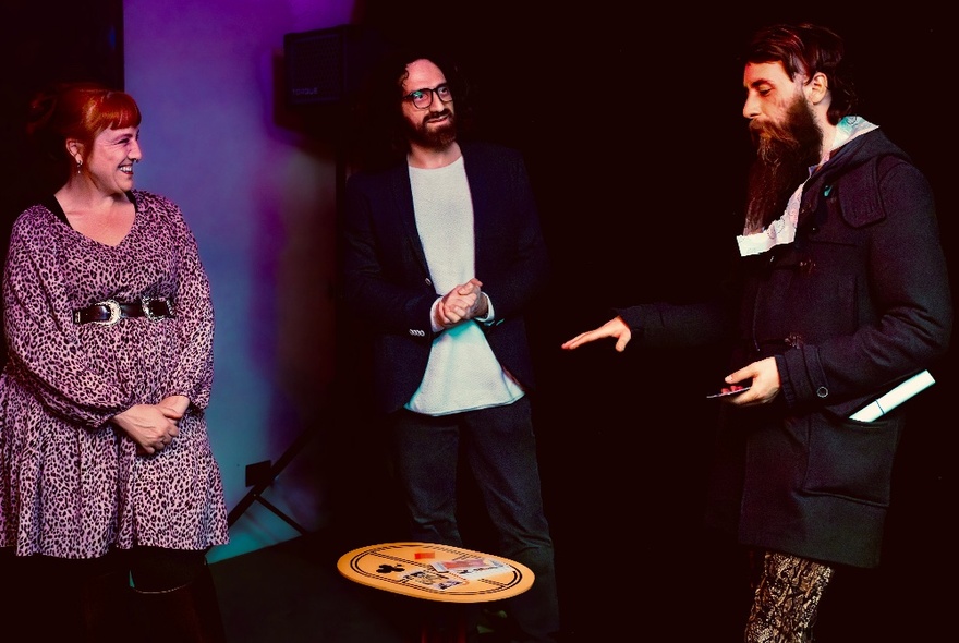 Three people stand around a small table while one performs a magic trick. 