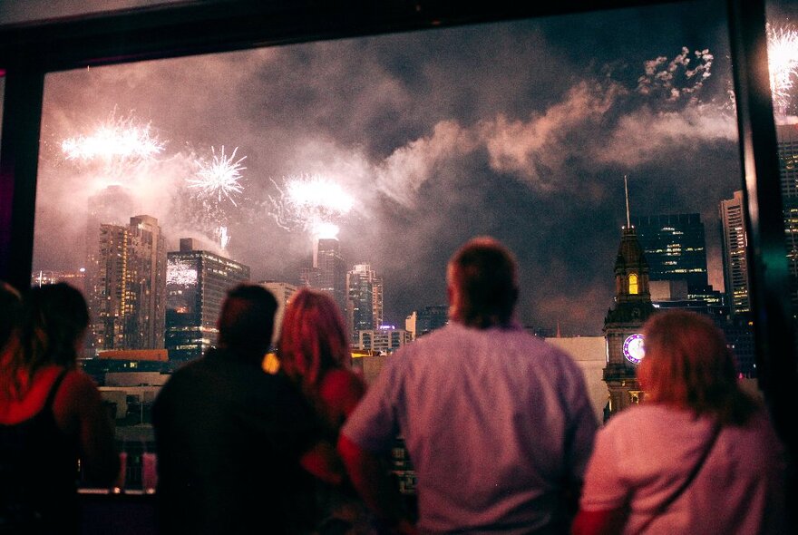 People gathered at The Stolen Gem Rooftop Bar watching fireworks over the city's buildings in the night sky.