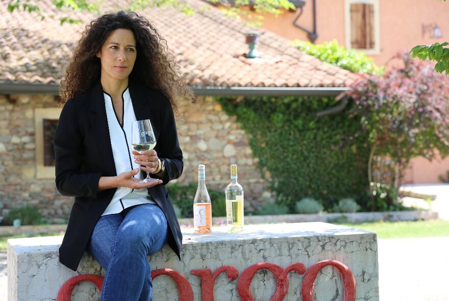 A woman holding a wine glass in her hand, seated on stone slab with two wine bottles next to her, in front of a country style chalet in the background.