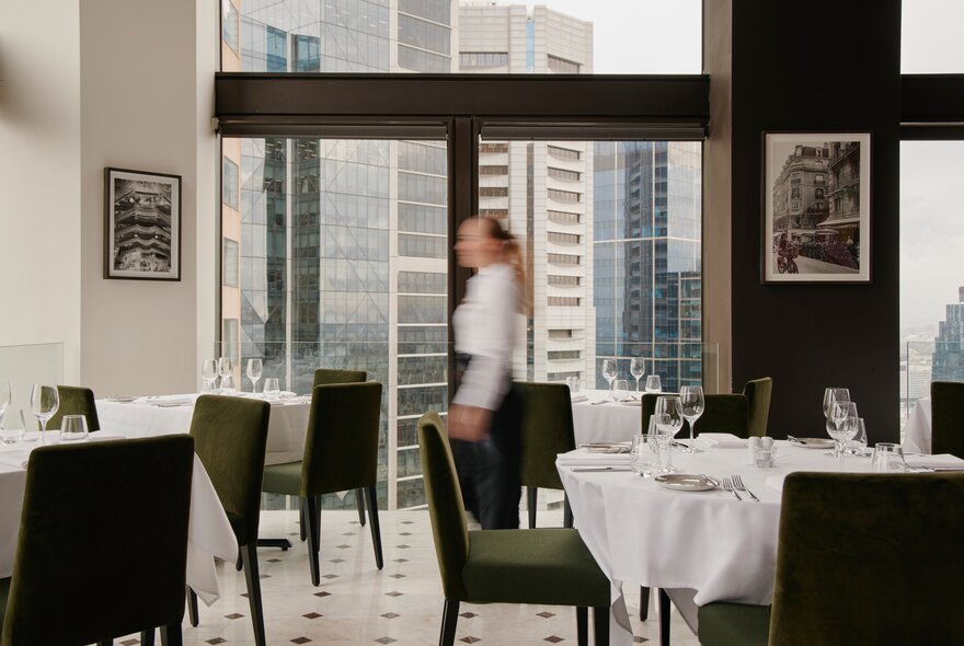 A blurred person walking between the dining tables and chairs in the No35 restaurant on the 35th floor of the Sofitel Melbourne with panoramic city views in the background.