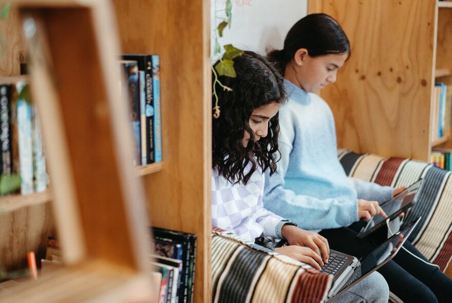 Two teenage girls working on their laptops, seated on a couch, with a bookshelf on either side of them.