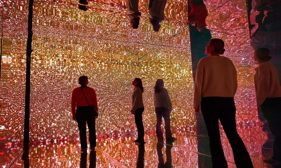 Three people standing in a colourful, sparkling exhibition with mirrors on the floor, walls and ceiling.