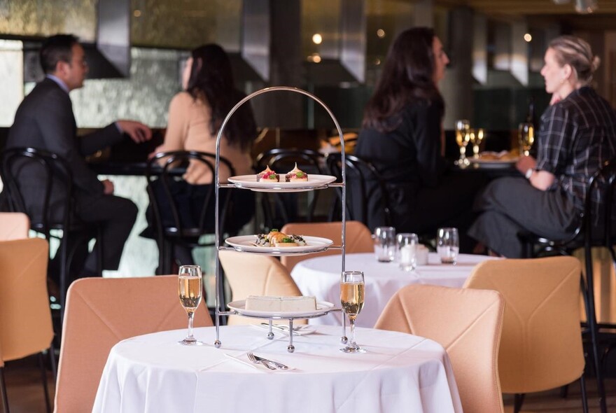 Customers sitting on stools behind a white napery table with three-tiered cake stand and glasses of bubbles.