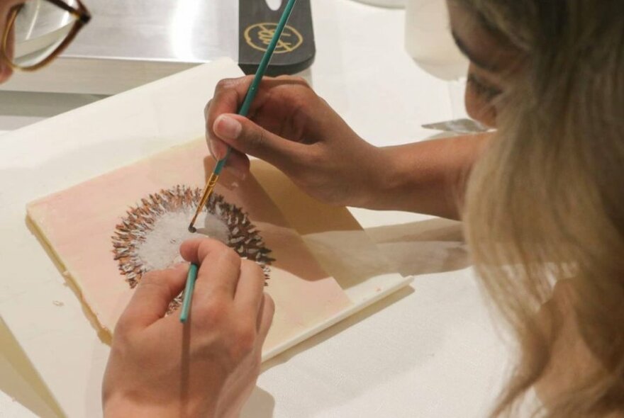 Hands using fine brushes to paint petals on a pink canvas of white chocolate.